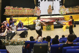 Traditional Indonesian dance performance on a decorated stage with gamelan musicians and two dancers in center, audience watching.