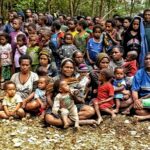 Large gathering of families, including many children, seated on the dirt ground in a forested area.