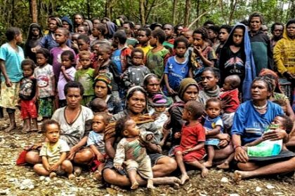 Large gathering of families, including many children, seated on the dirt ground in a forested area.