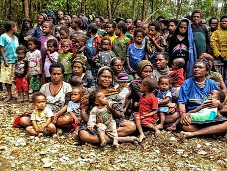 Large gathering of families, including many children, seated on the dirt ground in a forested area.