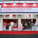 Wide storefront sign for KOPERASI DESA MERAH PUTIH, with a man in a suit on the right and shop posters visible inside the entrance.