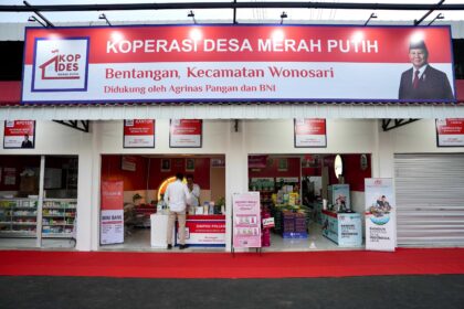 Wide storefront sign for KOPERASI DESA MERAH PUTIH, with a man in a suit on the right and shop posters visible inside the entrance.