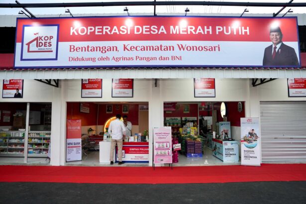Wide storefront sign for KOPERASI DESA MERAH PUTIH, with a man in a suit on the right and shop posters visible inside the entrance.