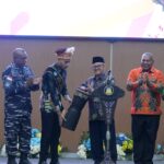 Group of men on a stage in colorful batik shirts clapping as a man with a feathered headpiece receives a ceremonial prop from a host at a formal event.