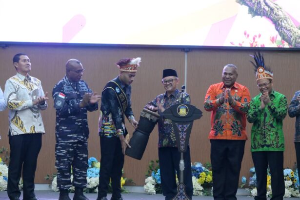 Group of men on a stage in colorful batik shirts clapping as a man with a feathered headpiece receives a ceremonial prop from a host at a formal event.