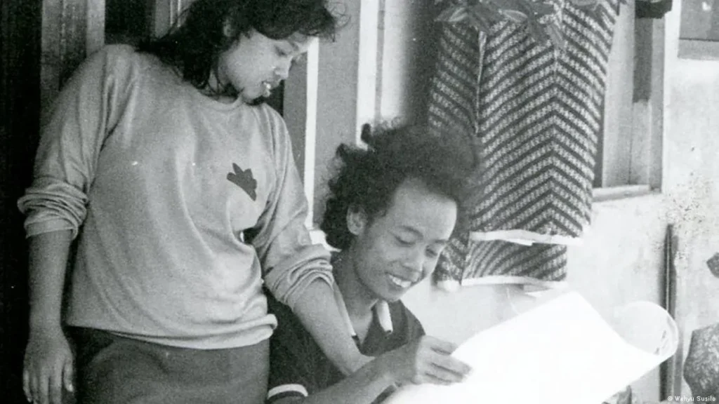 Two children outside a building lean over a large sheet of paper, looking at it together.
