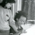 Two children outside a building lean over a large sheet of paper, looking at it together.