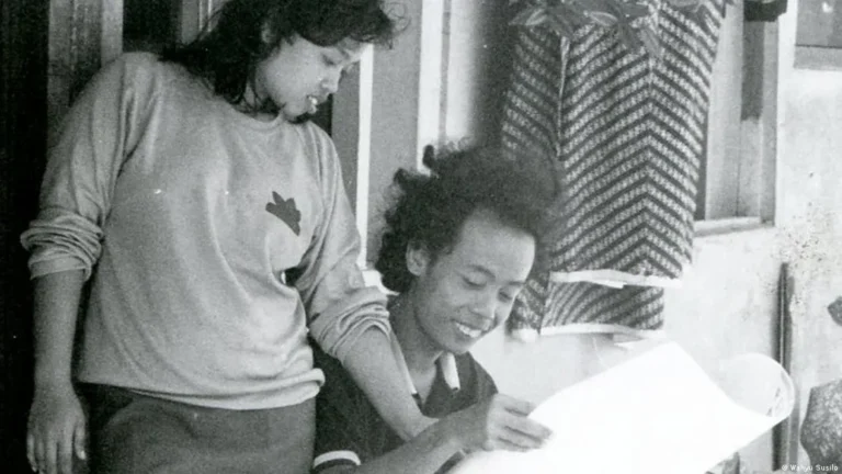 Two children outside a building lean over a large sheet of paper, looking at it together.