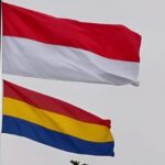 Two flags fluttering in the wind: the red-over-white Indonesian flag above a horizontal red-yellow-blue tricolor flag beneath it, against a pale sky.