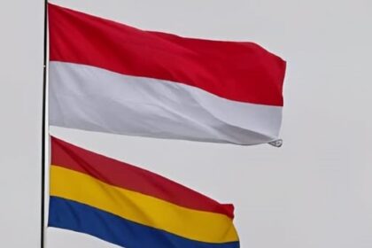 Two flags fluttering in the wind: the red-over-white Indonesian flag above a horizontal red-yellow-blue tricolor flag beneath it, against a pale sky.