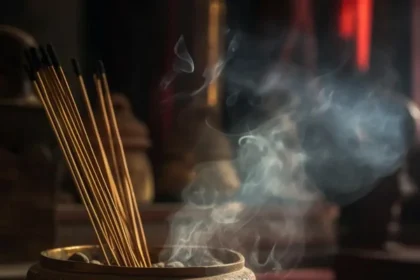 Incense sticks stand in a round wooden burner with wisps of smoke rising in a dimly lit room, creating a calm atmosphere.