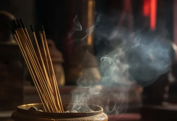 Incense sticks stand in a round wooden burner with wisps of smoke rising in a dimly lit room, creating a calm atmosphere.