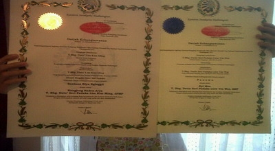 Two diplomas with green laurel borders are being held up by hands on each side, left featuring a gold seal and right a blue seal.