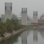 Construction site along a canal with tall concrete pillars and exposed steel reinforcements on top; a red crane visible in the background on a hazy day.