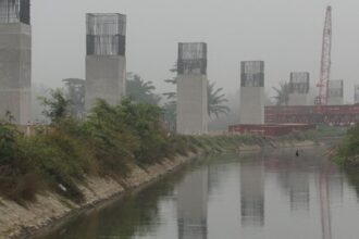 Construction site along a canal with tall concrete pillars and exposed steel reinforcements on top; a red crane visible in the background on a hazy day.