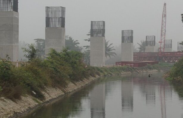 Construction site along a canal with tall concrete pillars and exposed steel reinforcements on top; a red crane visible in the background on a hazy day.