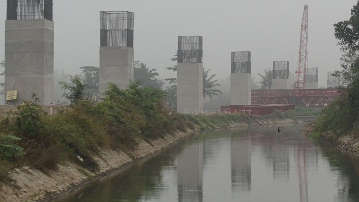 Construction site along a canal with tall concrete pillars and exposed steel reinforcements on top; a red crane visible in the background on a hazy day.