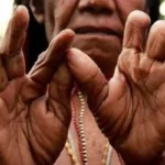 Close-up of dark-skinned hands forming a circle with thumbs and index fingers, beads visible in the background
