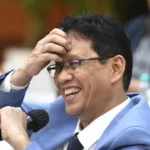 Man in light blue suit and glasses laughs while holding a microphone at a formal event.