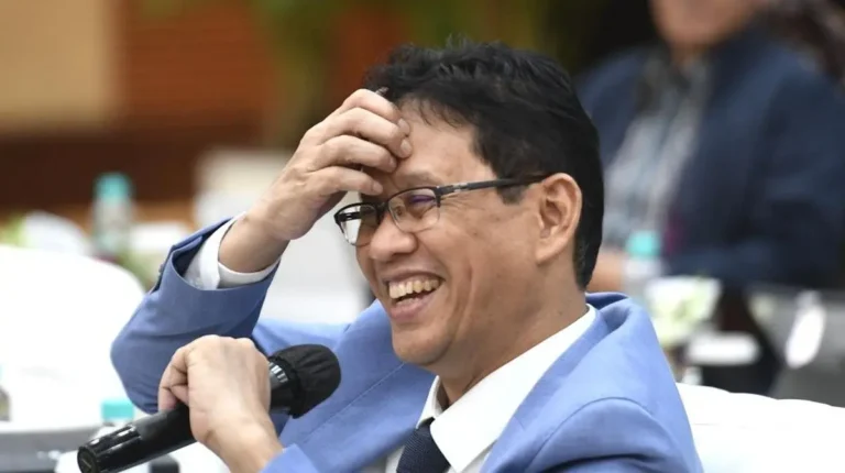 YRbl6kNaJ1 - Mabur.co Man in light blue suit and glasses laughs while holding a microphone at a formal event.