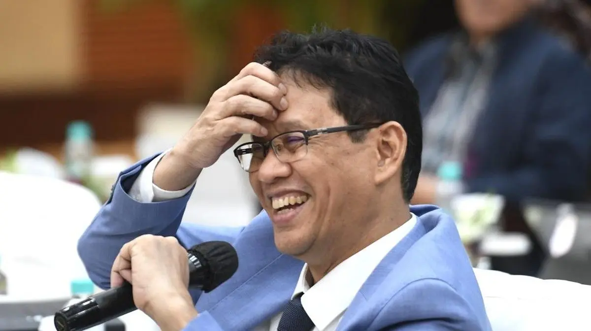 Man in light blue suit and glasses laughs while holding a microphone at a formal event.