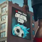 Billboard on a city building advertising YouTube FIFA Creator Cup with a blue-flamed soccer ball in the foreground Athlete’s leg visible for scale and action.
