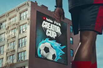 Billboard on a city building advertising YouTube FIFA Creator Cup with a blue-flamed soccer ball in the foreground Athlete’s leg visible for scale and action.