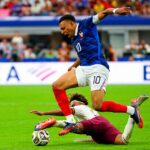 France footballer wearing blue jersey number 10 leaps over a sliding tackle toward the ball on a green pitch.