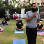 Instructor with microphone speaks to a group doing yoga outdoors on mats in a garden courtyard.