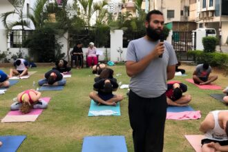 Instructor with microphone speaks to a group doing yoga outdoors on mats in a garden courtyard.
