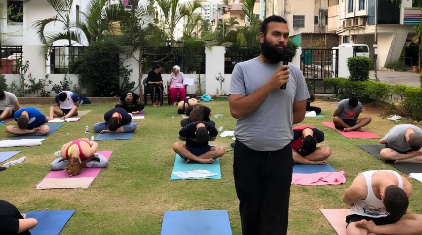 Instructor with microphone speaks to a group doing yoga outdoors on mats in a garden courtyard.