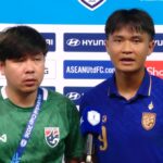 Two football players stand at a post-match press conference, one speaking into a blue microphone.
