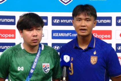 Two football players stand at a post-match press conference, one speaking into a blue microphone.