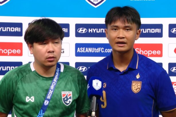 Two football players stand at a post-match press conference, one speaking into a blue microphone.