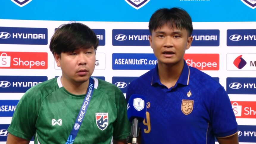 Two football players stand at a post-match press conference, one speaking into a blue microphone.