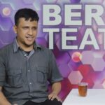 Man in a gray shirt sits beside a white board in a colorful studio backdrop that reads 'BERITA TEATER'. Two glasses of tea sit on a white table in front of him.