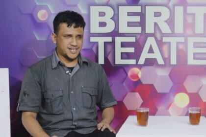 Man in a gray shirt sits beside a white board in a colorful studio backdrop that reads 'BERITA TEATER'. Two glasses of tea sit on a white table in front of him.