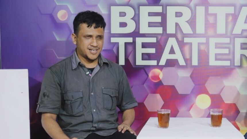 Man in a gray shirt sits beside a white board in a colorful studio backdrop that reads 'BERITA TEATER'. Two glasses of tea sit on a white table in front of him.