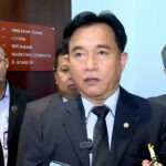Man in a dark suit and tie speaks to reporters, microphones in view, flanked by colleagues.