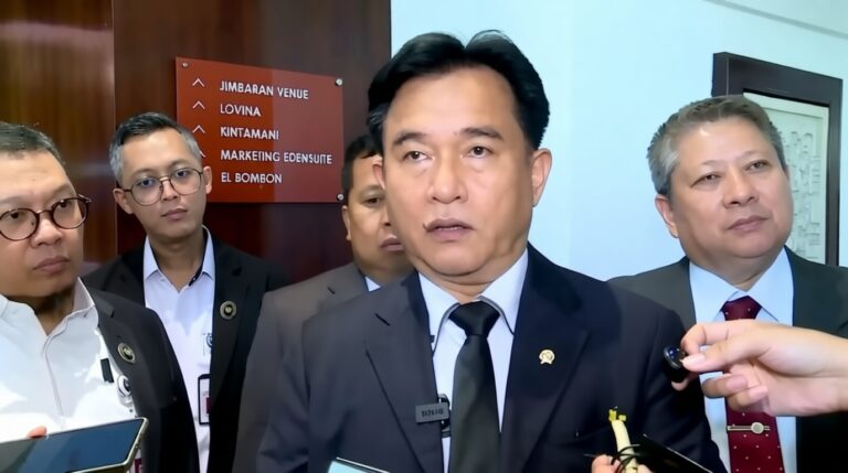 Man in a dark suit and tie speaks to reporters, microphones in view, flanked by colleagues.