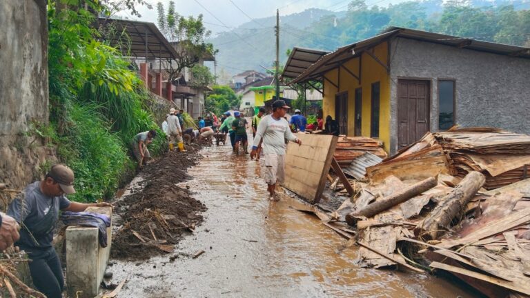 A flooded village street where people are shoveling mud and removing debris together after a flood (community cleanup).