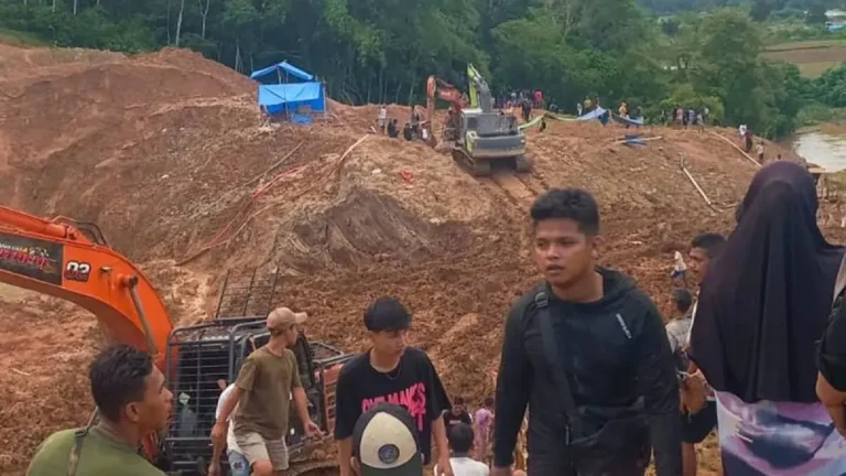 =a - Mabur.co Construction site on a muddy slope with an orange excavator and workers; crowd visible in the background on the hill carven by earthworks.