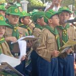 Group of young scouts in khaki uniforms with green hats and scarves, holding booklets during a parade. One child sings into a microphone while others watch.