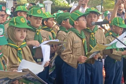 Group of young scouts in khaki uniforms with green hats and scarves, holding booklets during a parade. One child sings into a microphone while others watch.