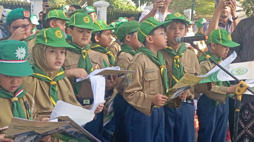 Group of young scouts in khaki uniforms with green hats and scarves, holding booklets during a parade. One child sings into a microphone while others watch.