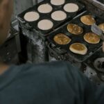 Cook flips pancakes on a commercial griddle with round molds, several pancakes cooking on both sides.