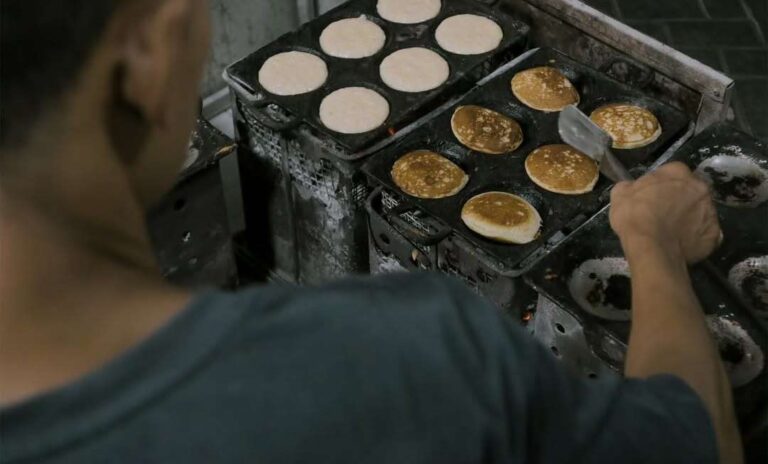 Cook flips pancakes on a commercial griddle with round molds, several pancakes cooking on both sides.