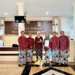 Group of six people in matching red traditional outfits stand in a government office lobby, one woman in green hijab holds a document while another holds a decorative basket and gift.