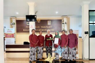 Group of six people in matching red traditional outfits stand in a government office lobby, one woman in green hijab holds a document while another holds a decorative basket and gift.