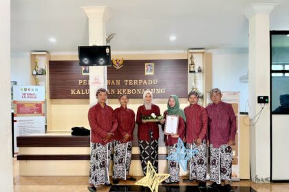 Group of six people in matching red traditional outfits stand in a government office lobby, one woman in green hijab holds a document while another holds a decorative basket and gift.